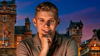 A young man in a brown shirt holds a hand to his chin as he smiles to camera, set against the backdrop of a castle by night