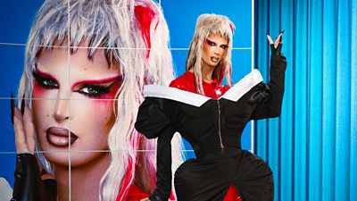 A drag queen in a mullet style wig (The Only Naomy) poses in red-tinged wig and exaggerated 80s-style off-shoulder dress 