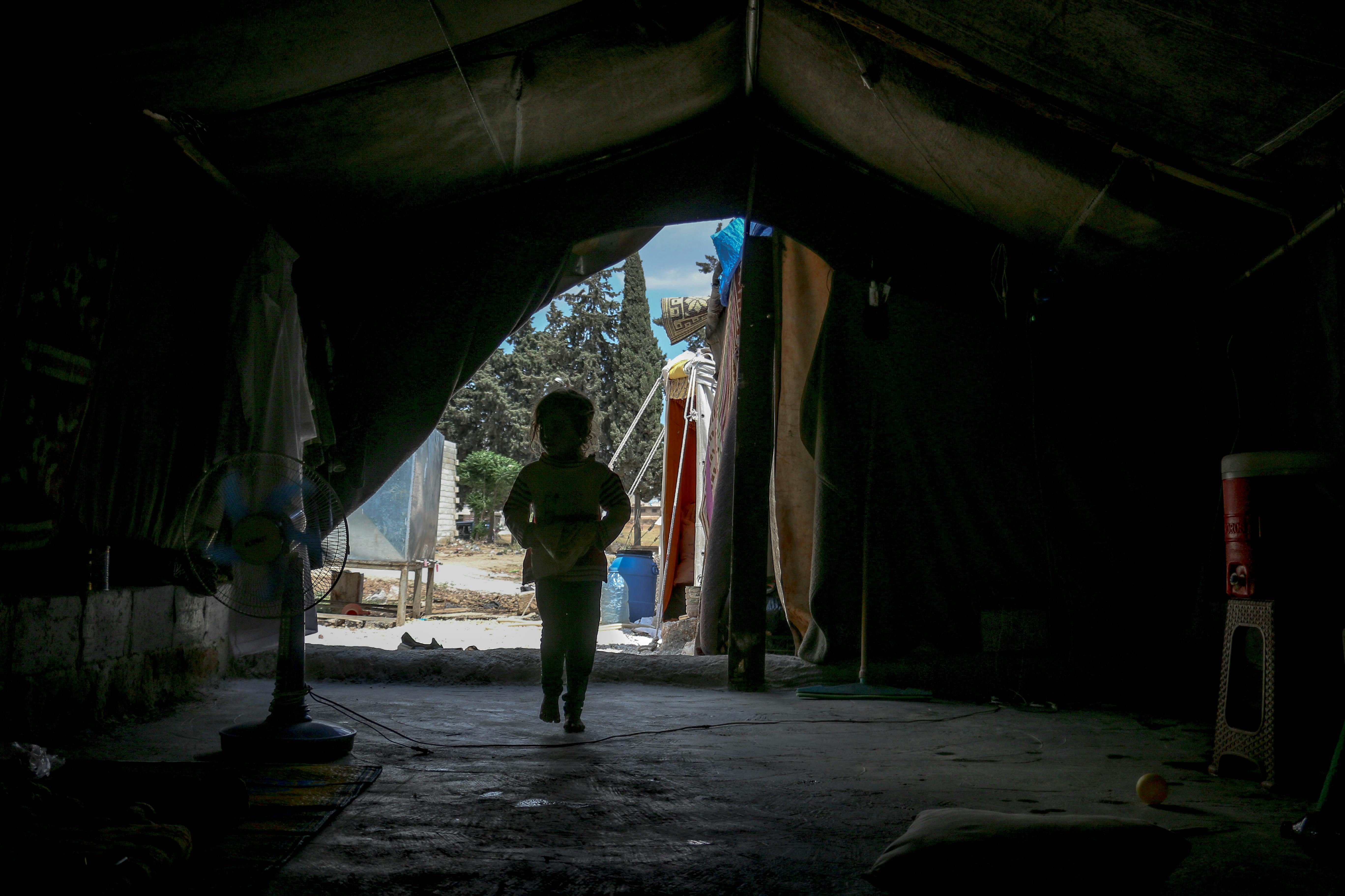 Silhouette of a young child standing inside a tent, looking out toward daylight and trees outside.