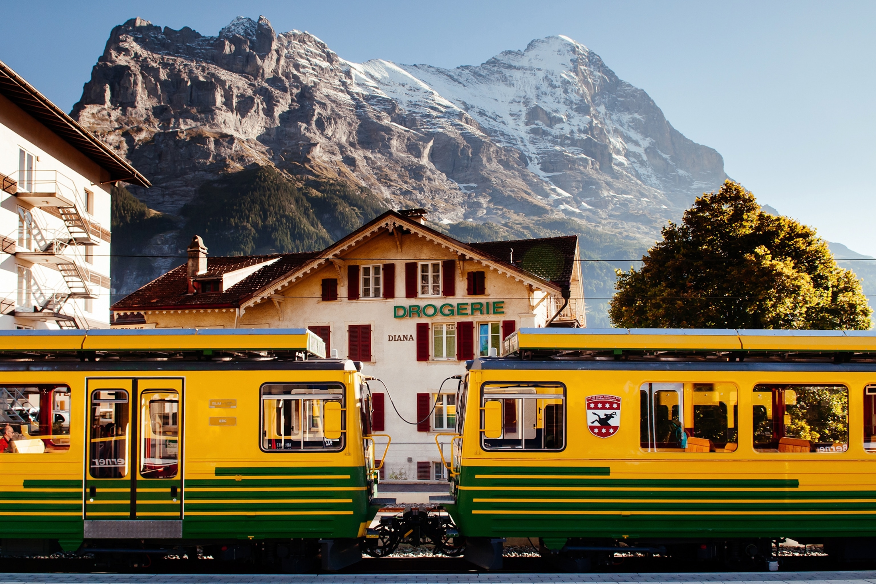 A kitsch train stopping at a mountain station in the Swiss Alps with snow-capped mountains in the background.