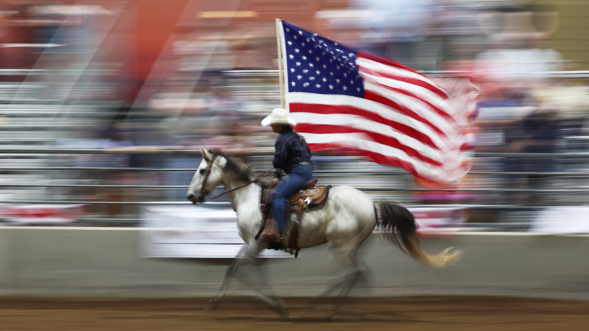 A cowboy on a horse kicks off a rodeo by riding around an arena with a US flag.