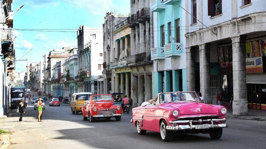 Old cars drive through a street in Havana, Cuba.