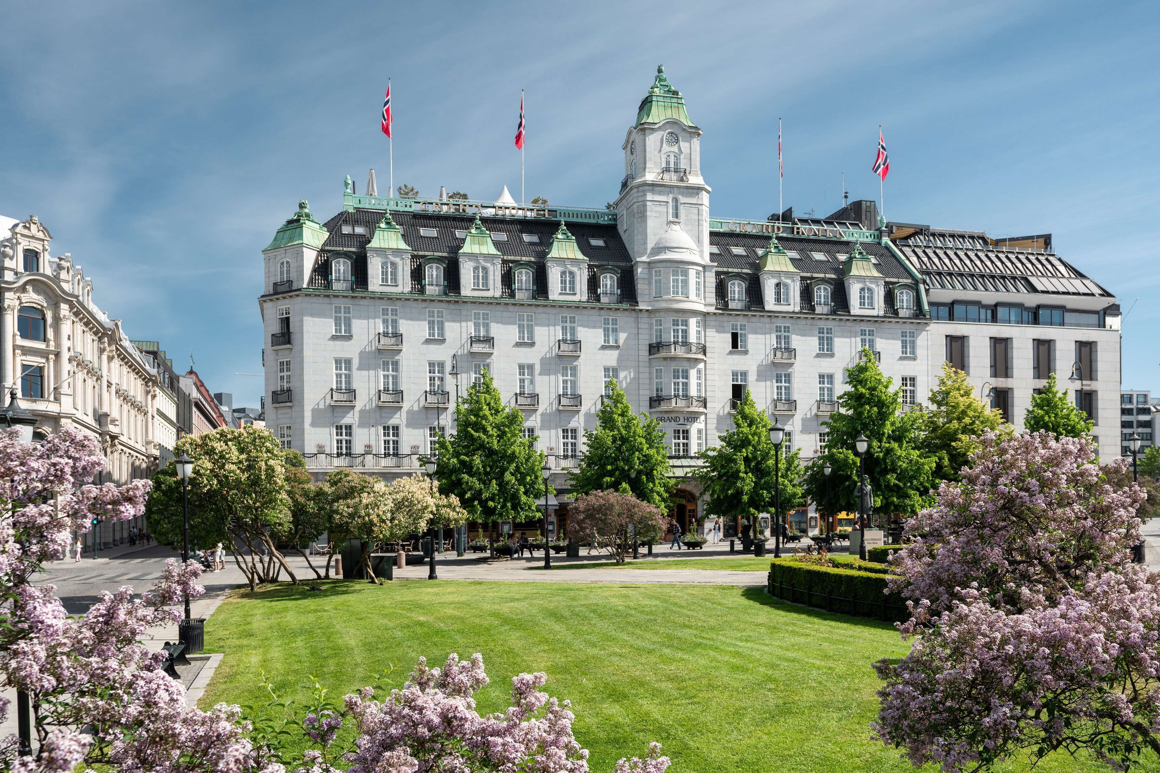 The Grand Hotel Oslo, a white building with a green roof and tower, viewed from a park with green grass and blooming trees.
