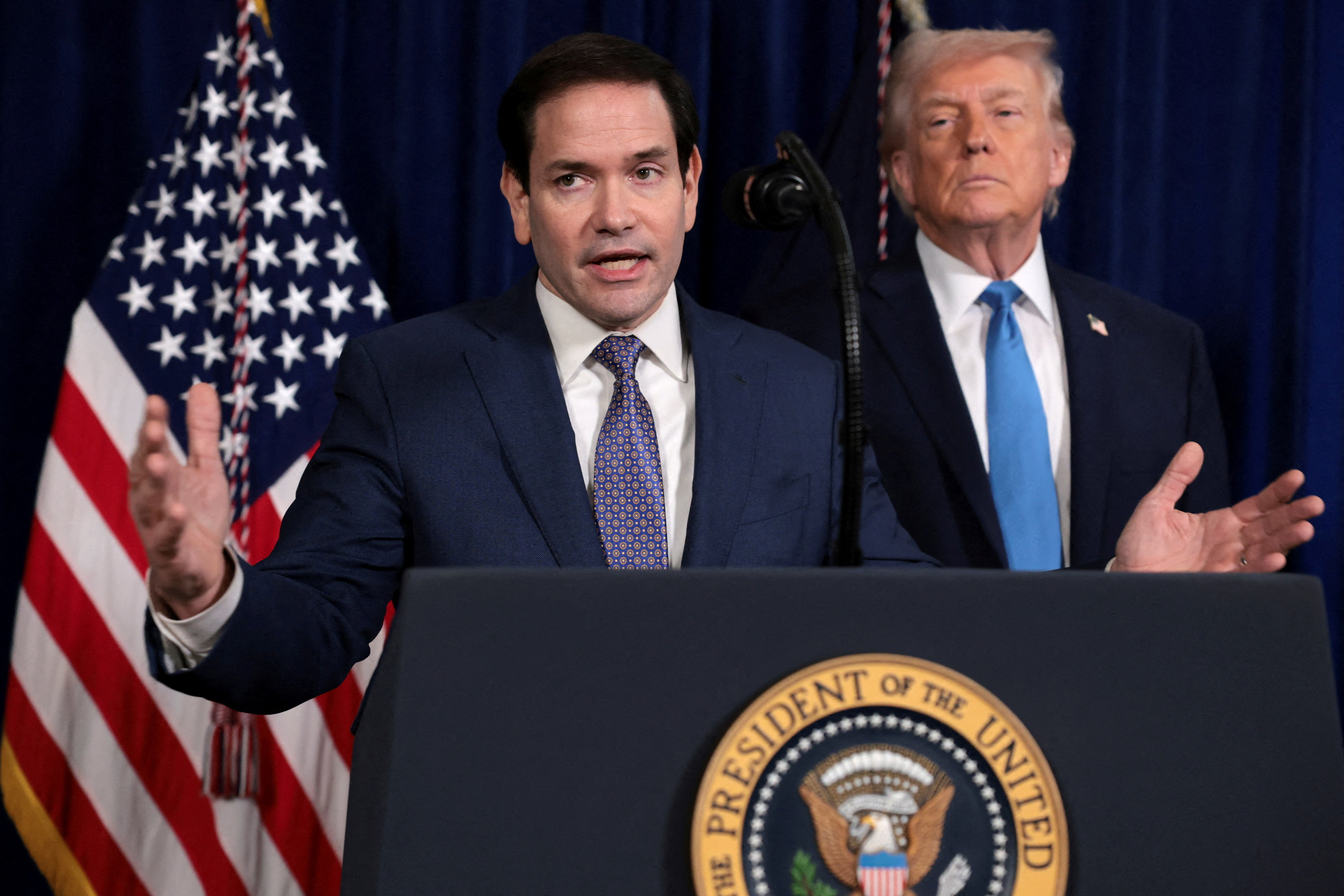 Marco Rubio speaking at a podium with Donald Trump behind him, with an American flag to the left.