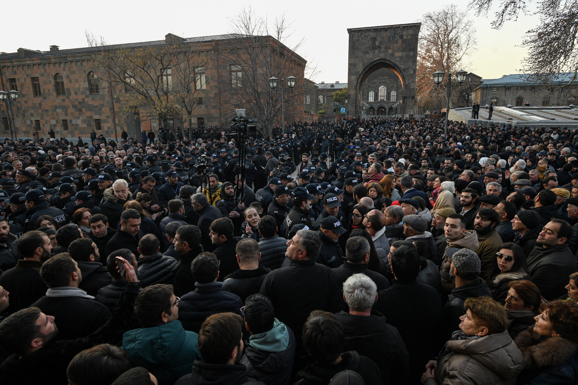 A large crowd of people, including many police officers, gathered in a square in front of a stone building with an arched entrance.