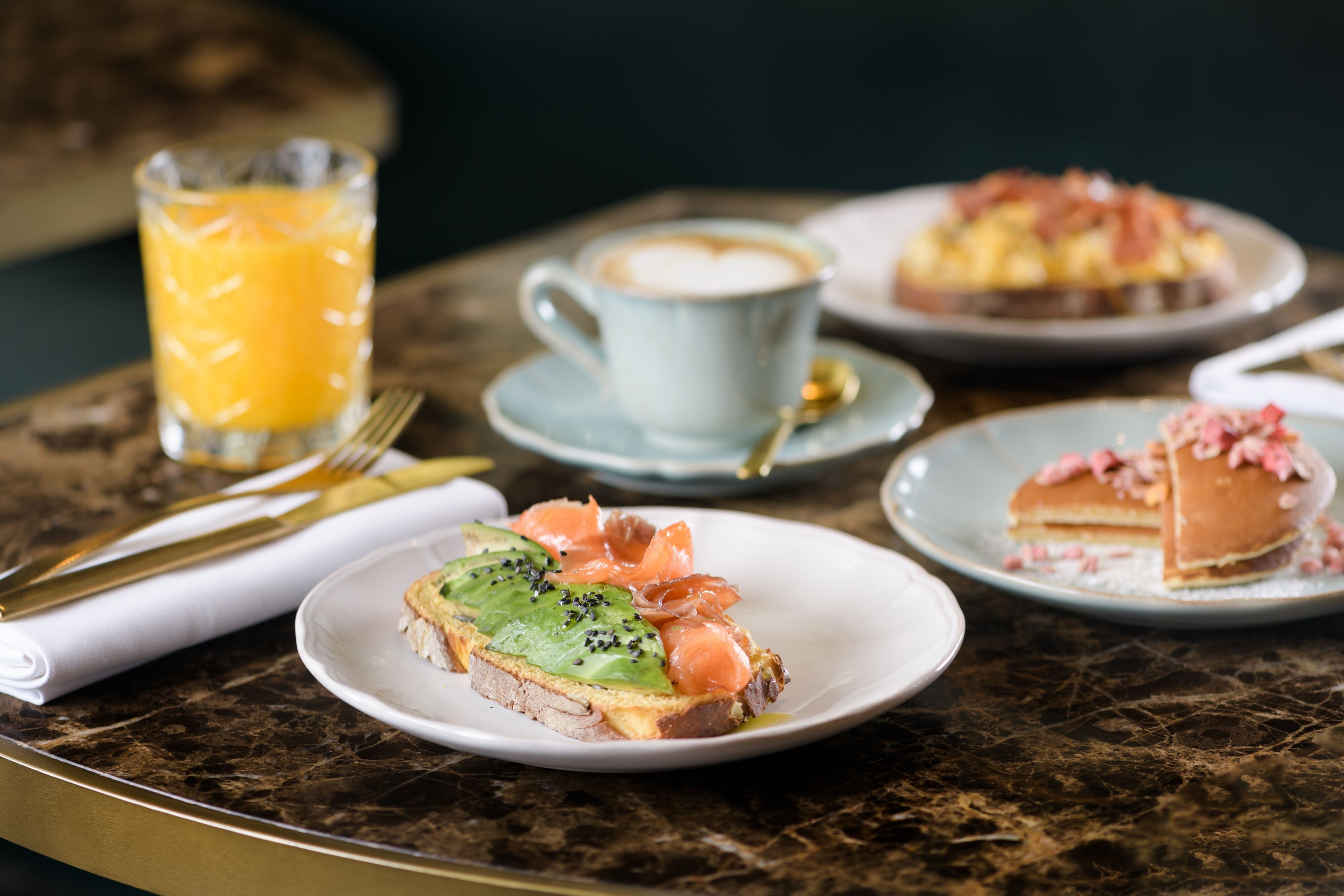 Breakfast spread with avocado and salmon toast, pancakes, and scrambled eggs, orange juice, and a cup of coffee.