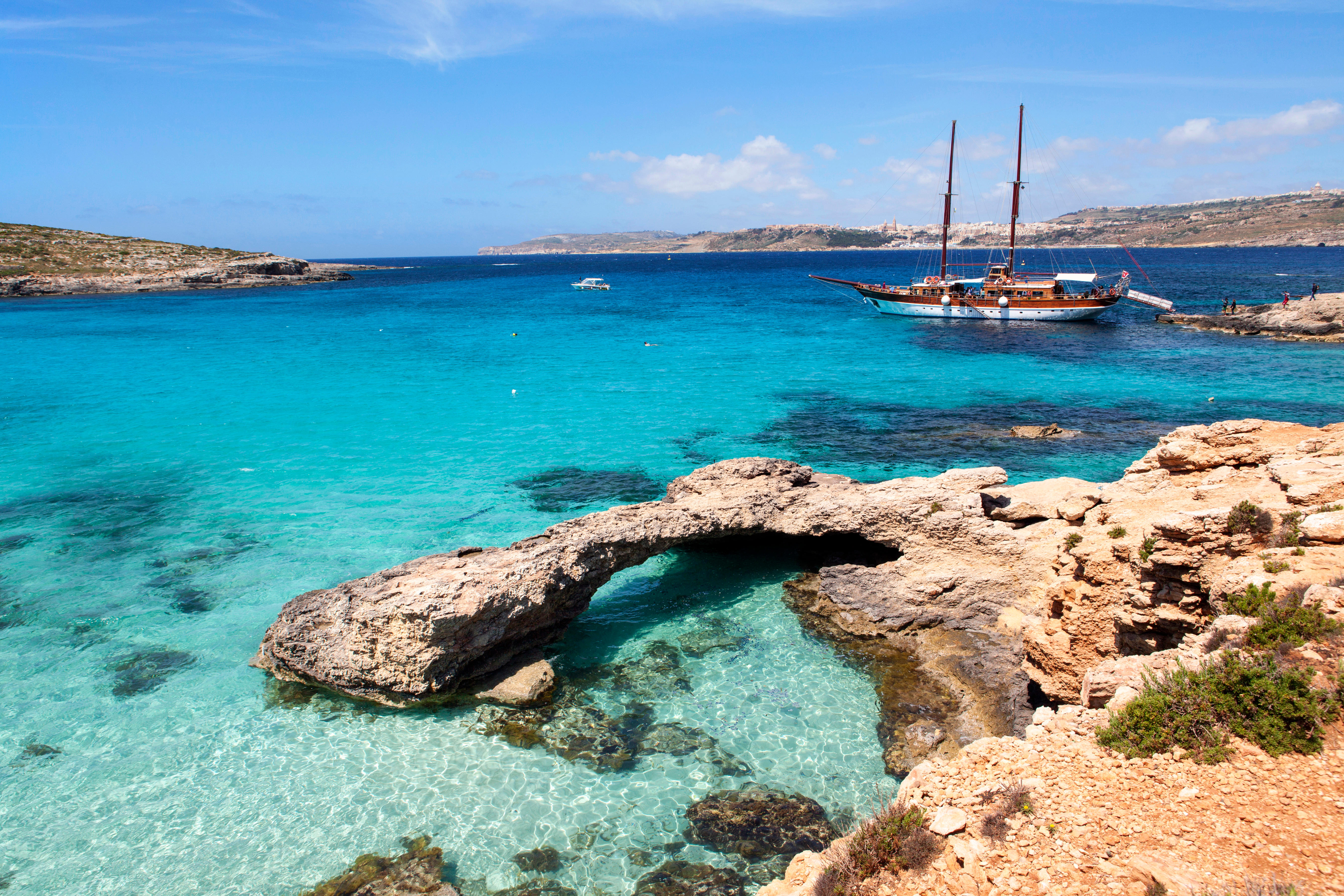 Blue Lagoon on Comino Island, Malta, with a boat anchored near the rocky shore and a natural stone arch over clear turquoise water.