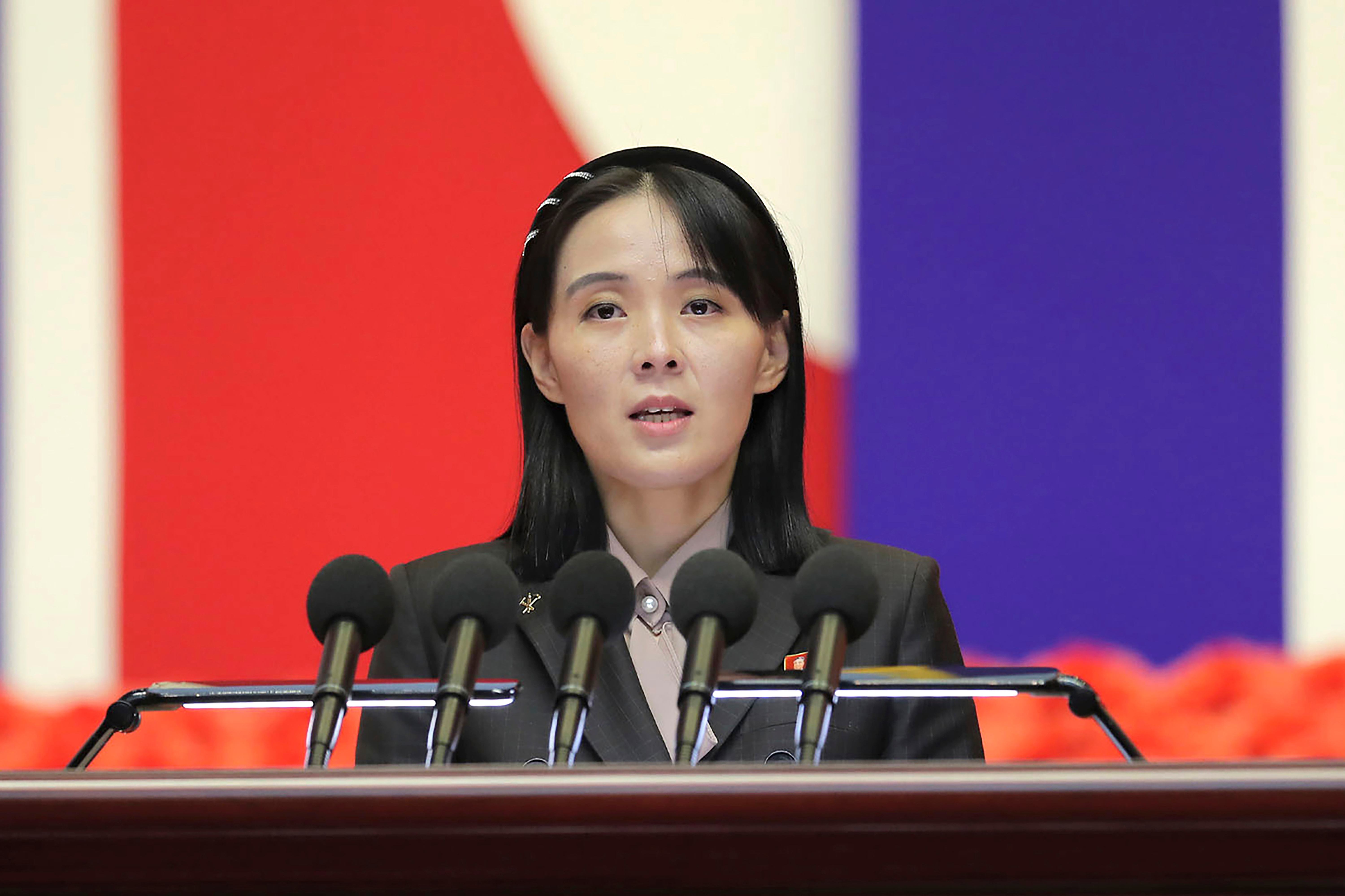 Kim Yo Jong speaking at a podium with microphones, with a red and blue background.