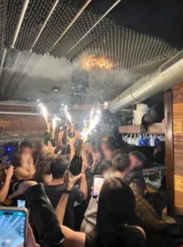 Fire igniting on the ceiling of a bar from sparkling champagne bottles held by blurred figures in a crowd.