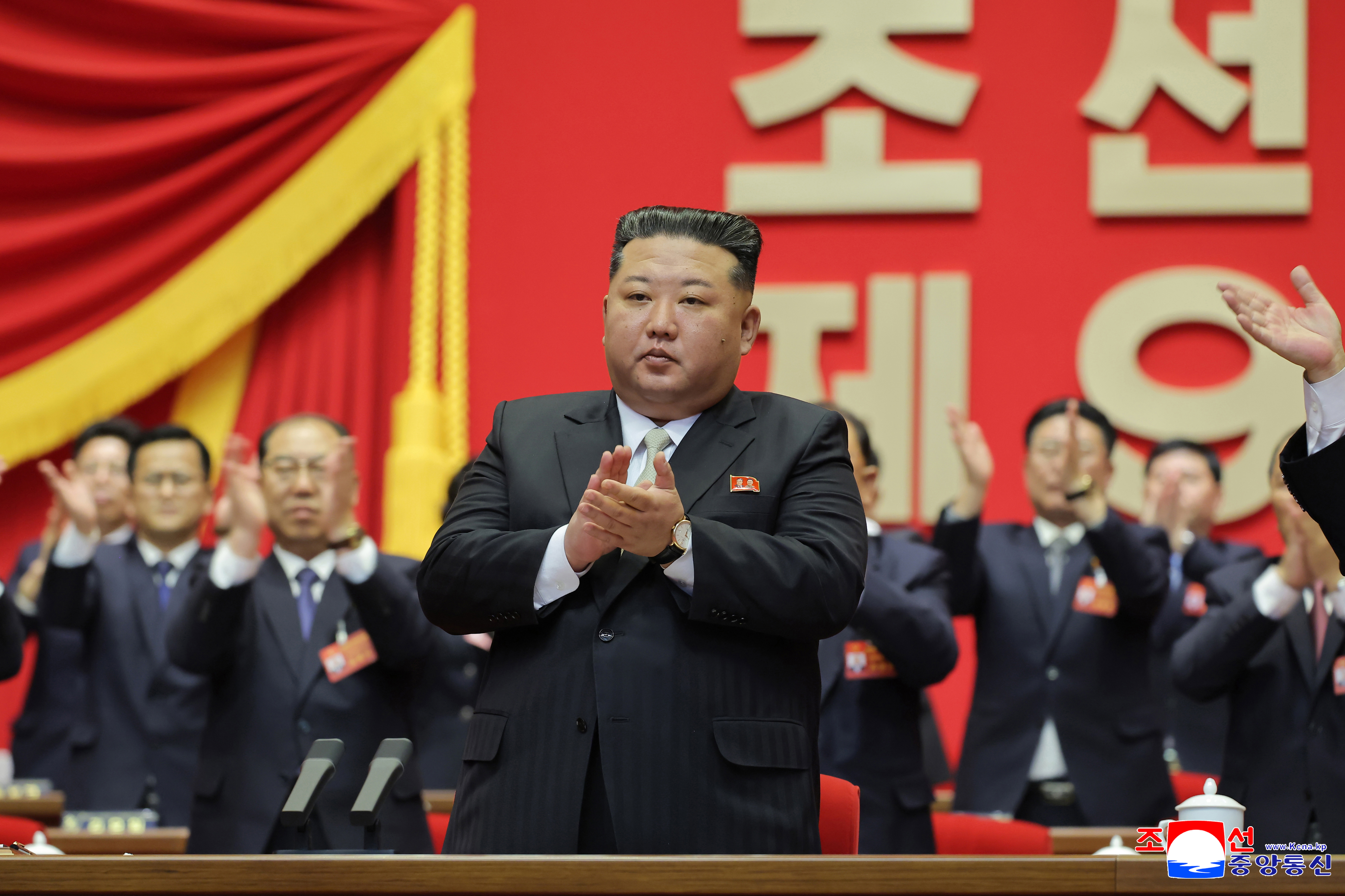 North Korean leader Kim Jong Un, in a dark suit, stands and applauds at a political congress, with other officials clapping in the background.
