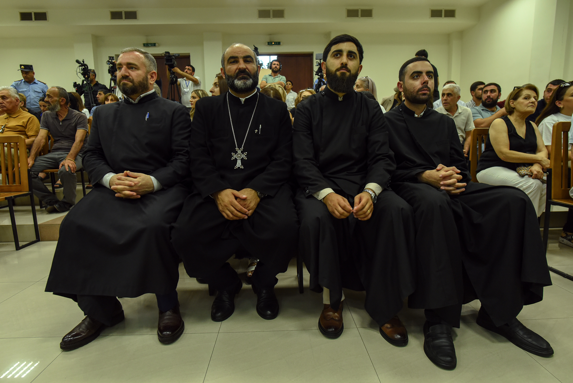Four men in black clerical attire sitting in a row, with other people and camera crew in the background.