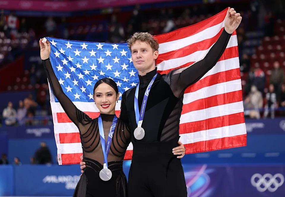 Two figure skaters in stylish black outfits hold an American flag and wear silver medals, standing on an ice rink at a competition