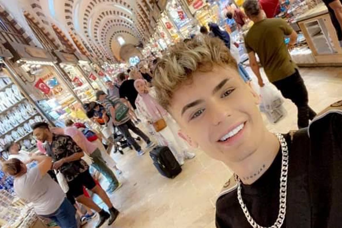 An individual with short, light-colored hair, wearing a black shirt and a necklace, is smiling while standing in a busy indoor shopping area with numerous people walking around.