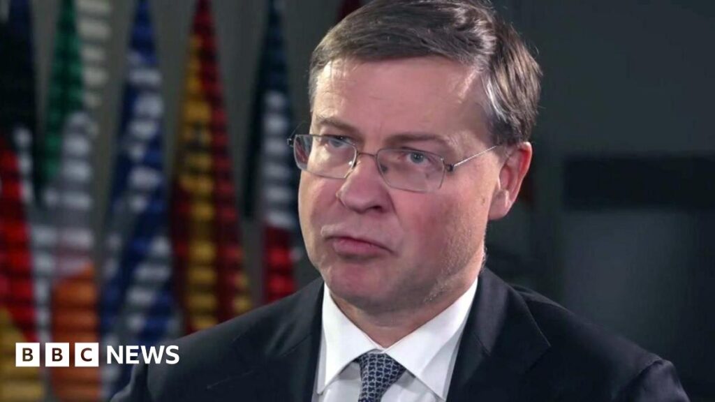 Valdis is wearing a suit and a patterned tie. He is sitting in a room and behind him is a row of colourful national flags displayed in the background.