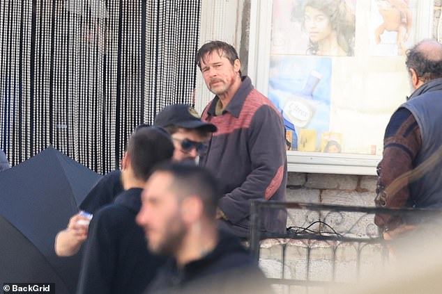 The American star looked in pain as he pulled a number of faces with blood dripping down his cheek before perched on a wall looking somber as a crowd of people walked past him
