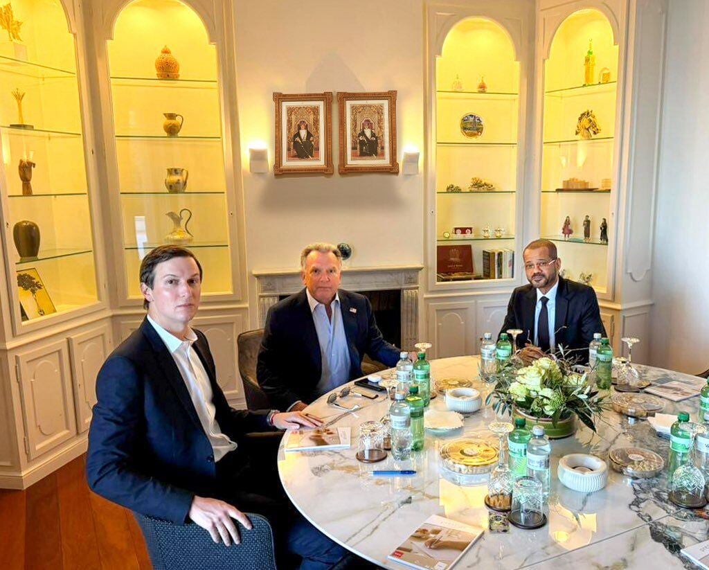 Three men are shown facing the camera while seated at a large circular table with glassware. The men are wearing suit jackets.
