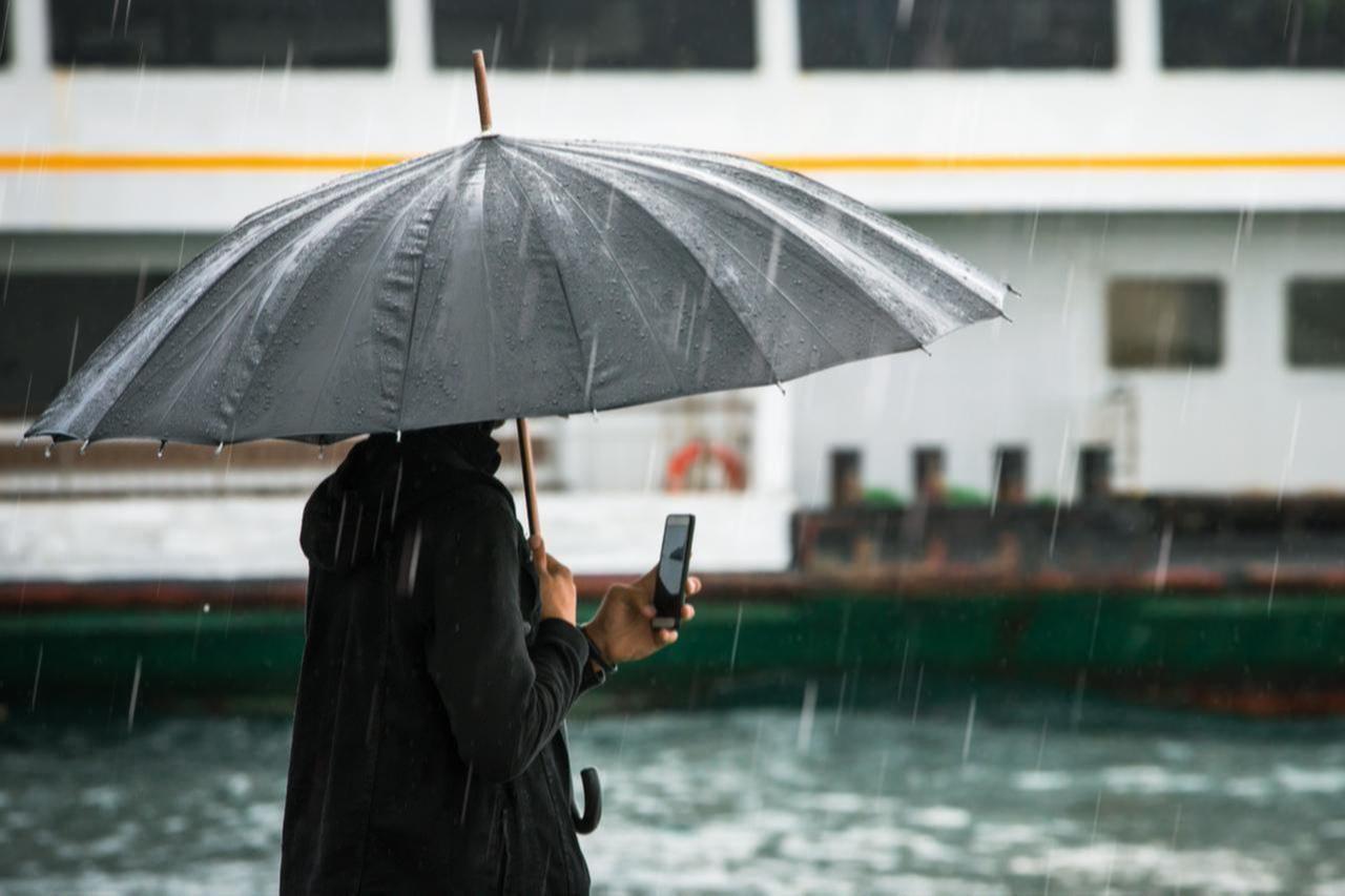 A person holds an umbrella while checking their phone near the waterfront during heavy rain in Istanbul, Türkiye. (Adobe Stock Photo)