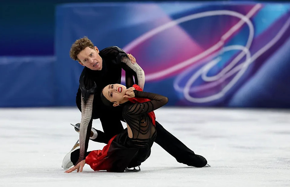 Two figure skaters perform an emotional routine on ice, wearing elegant black and red costumes
