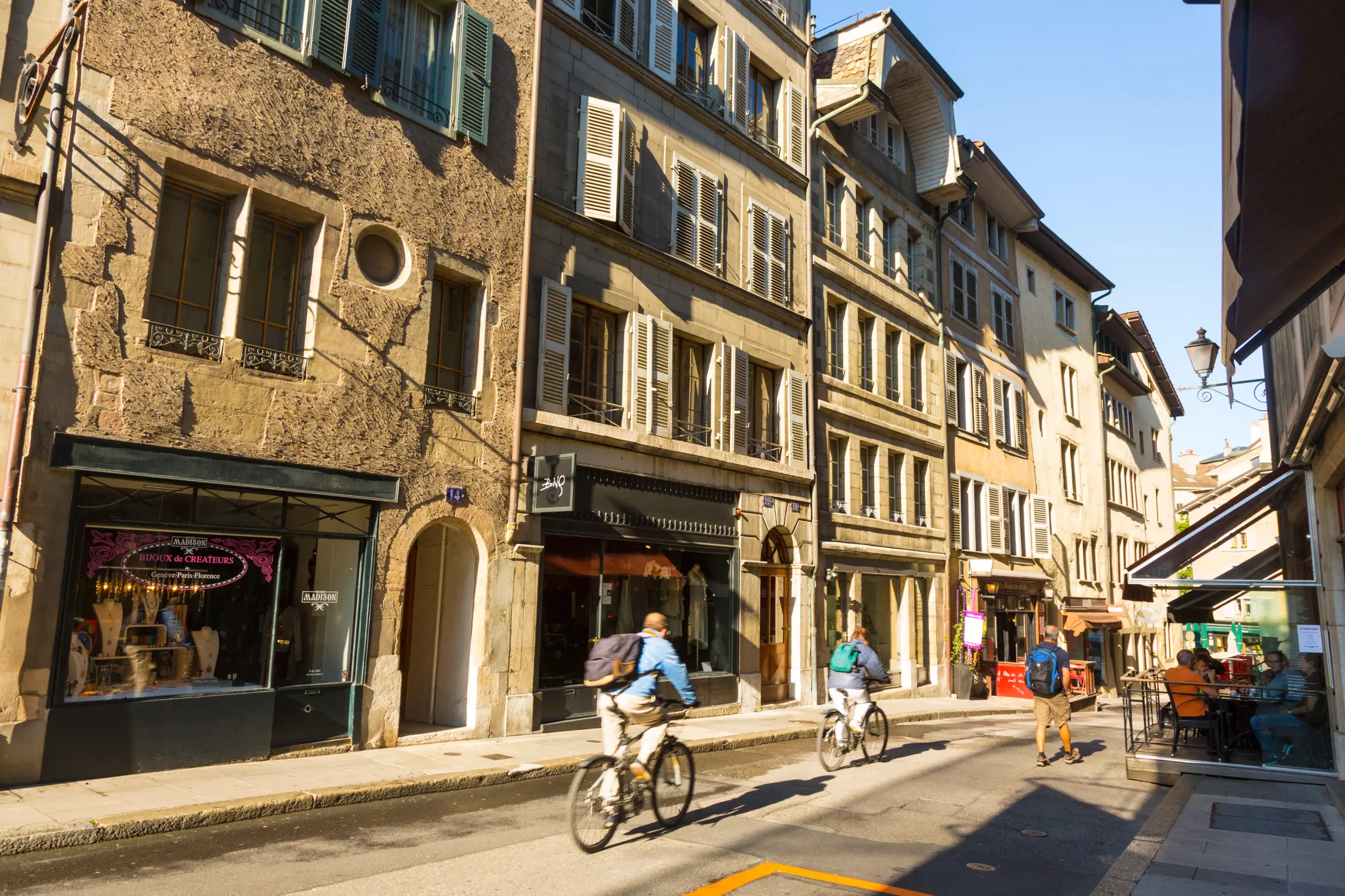 Cyclists and pedestrians moving along a sunny street in Old Town, Geneva, Switzerland, with buildings and shops lining the road.