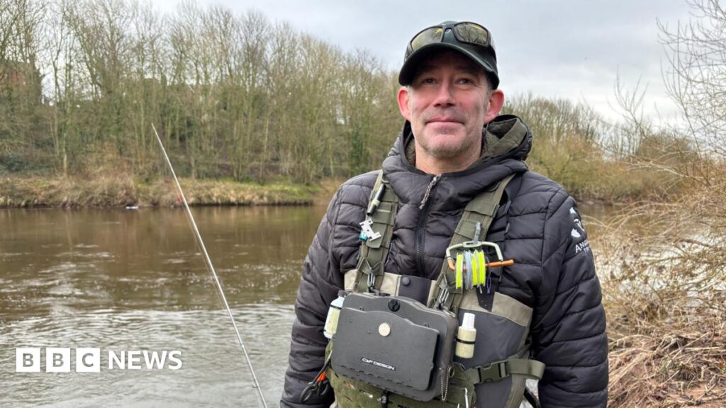 Del Spry standing in front of a river. He is wearing a black jacket, cap and sunglasses on his head and a khaki-coloured chest pack with fishing gear attached to it. The riverbank on the opposite side is lined with bare trees.