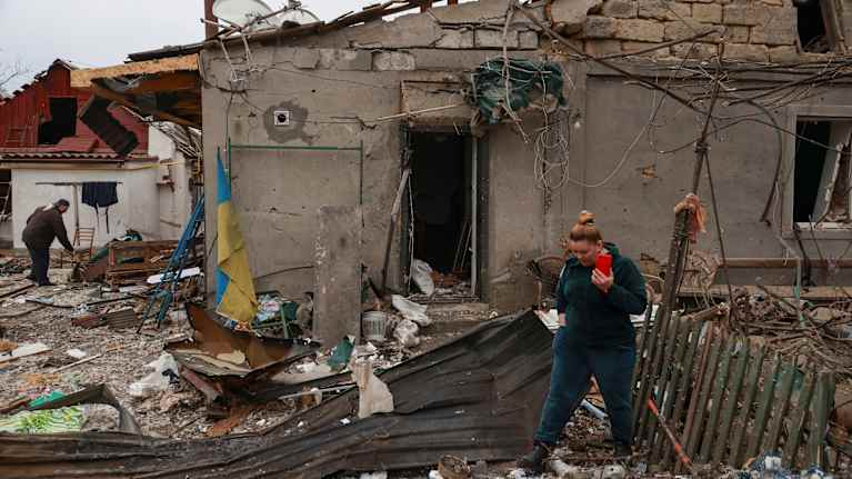Woman in green pants and top holding a smartphone walking in front of the remains of a building damaged in an air strike.