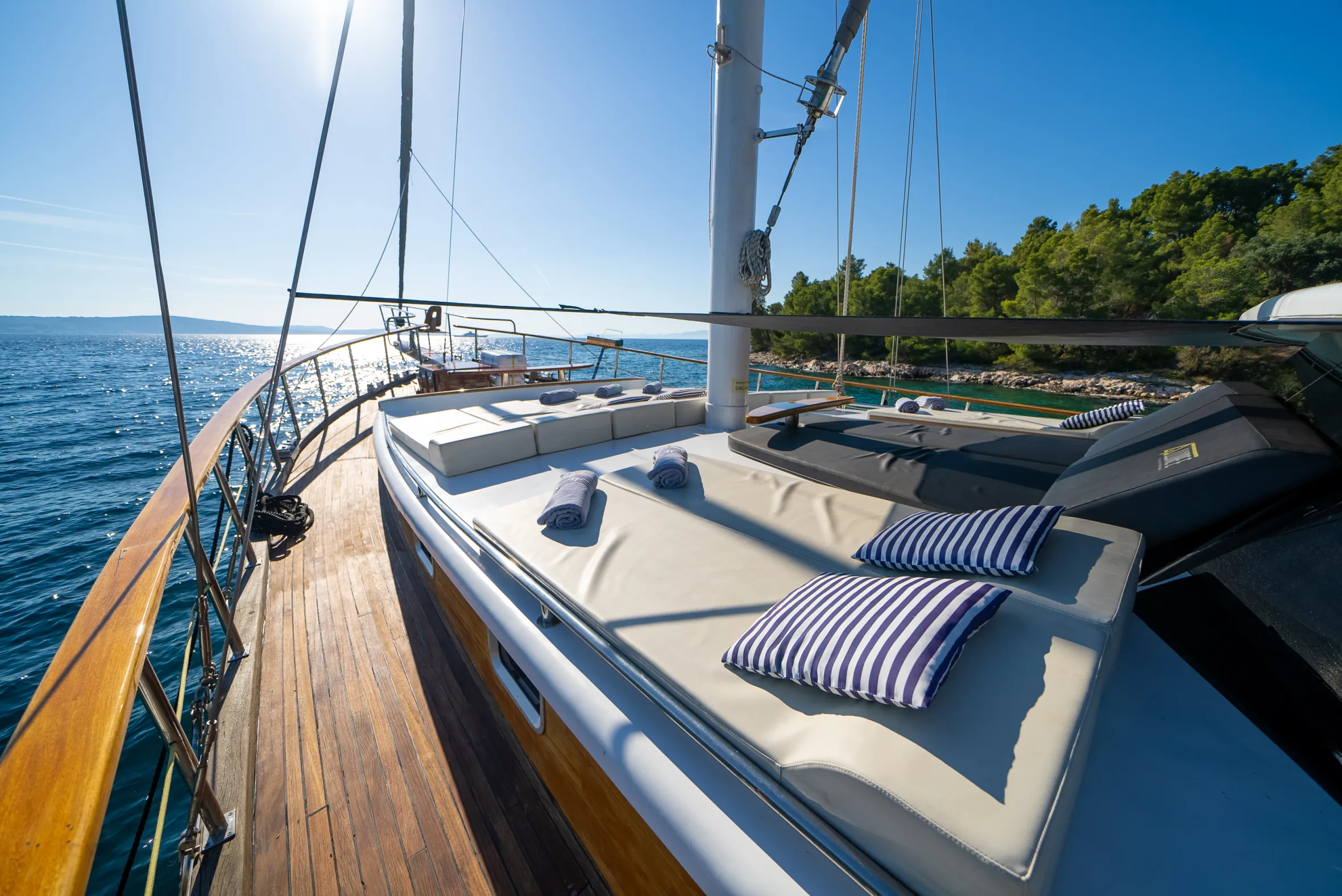 Deck of a sailing yacht with lounge seating overlooking the clear blue sea and a green coastline.