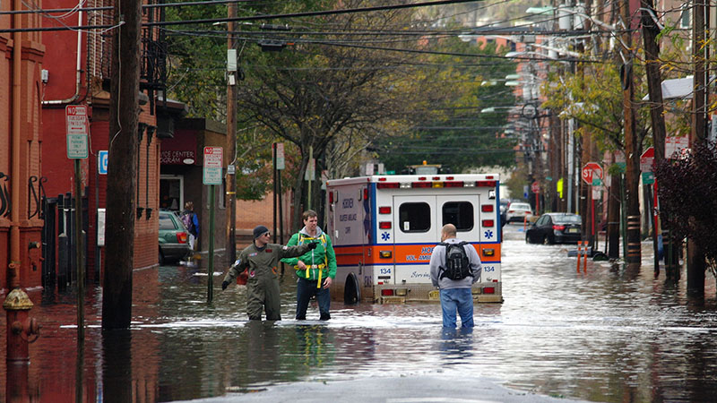 A flooded street with an emergency vehicles, power lines, and people wading through the water.