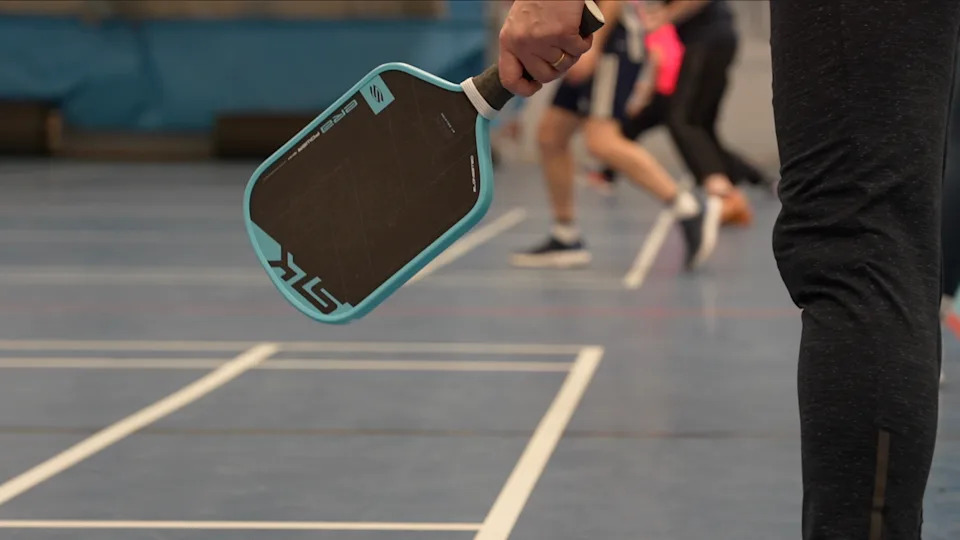 A hand holding a pickleball paddle, face open. The paddle has a light blue rim, and a black surface where the polymer ball strikes. There are players, mid-match, in the background. 