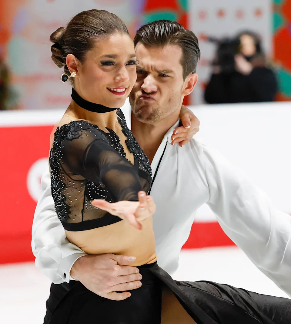 Two figure skaters perform a routine on the ice. The woman wears a sheer, embellished top with a long skirt; the man wears a simple white shirt