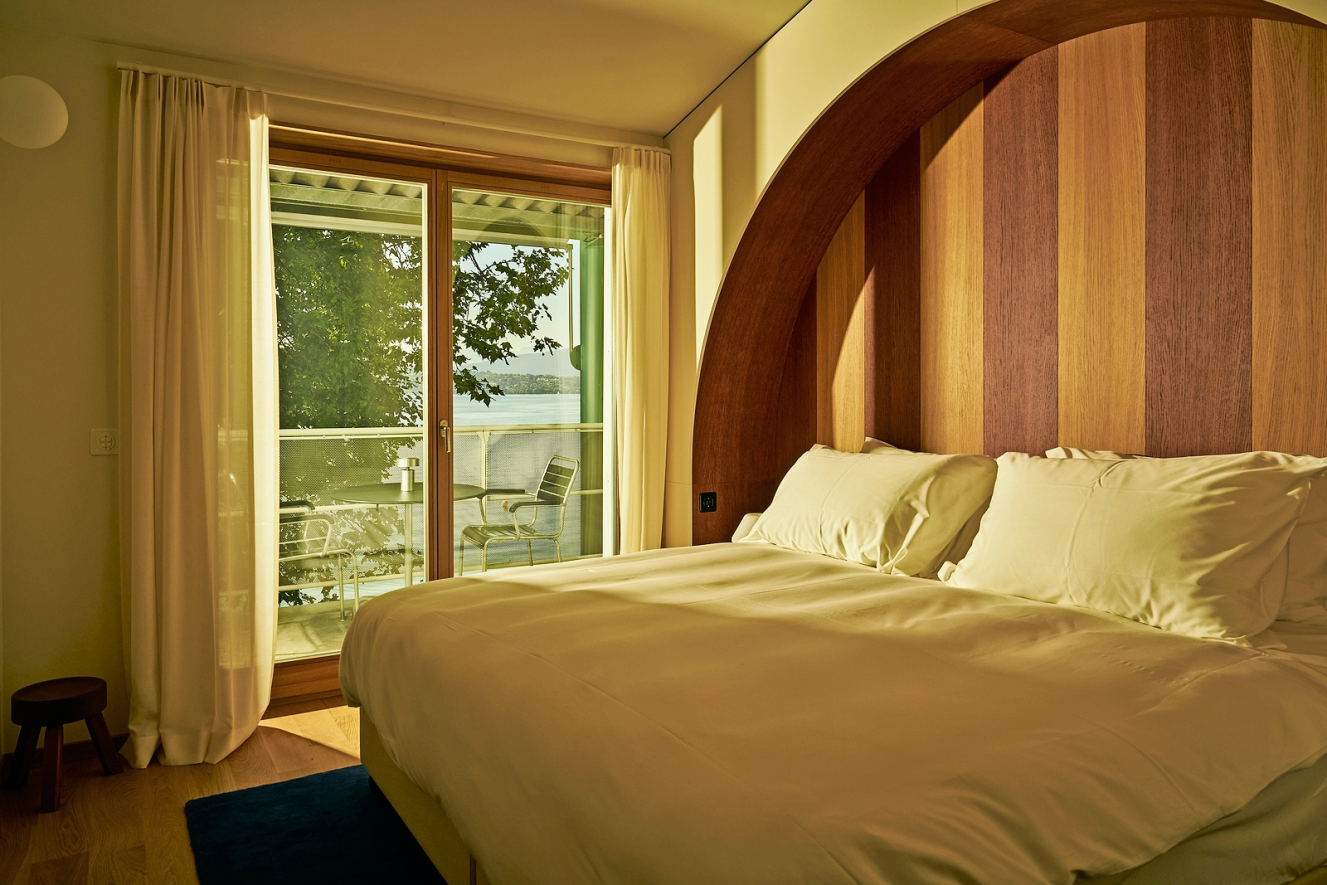 A bedroom at Auberge Port-Gitana in Geneva, Switzerland, with a bed, a wooden headboard, and a balcony overlooking a lake.