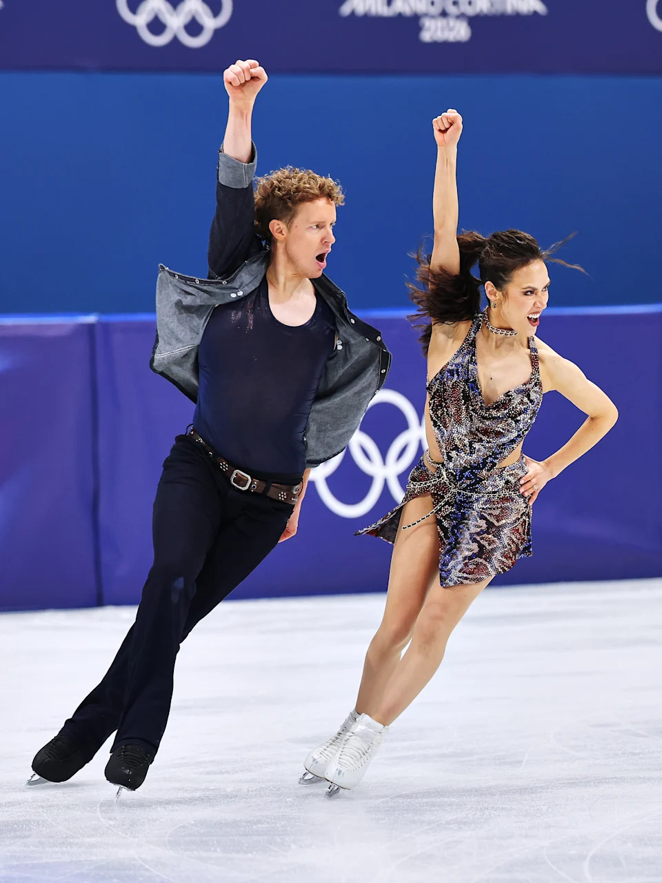 Two figure skaters perform energetically on ice at the Olympics. The man wears a denim jacket, and the woman wears a sequin dress