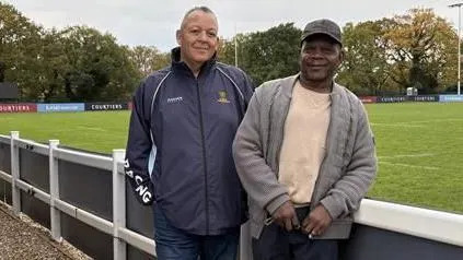 Two men stand side by side smiling into the camera. Both are wearing waterproof style jackets and the man on the right has a dark baseball cap on his head.
