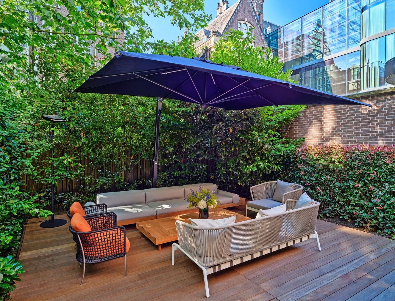 An outdoor seating area with sofas, chairs, a coffee table, and a large umbrella, surrounded by lush green foliage.
