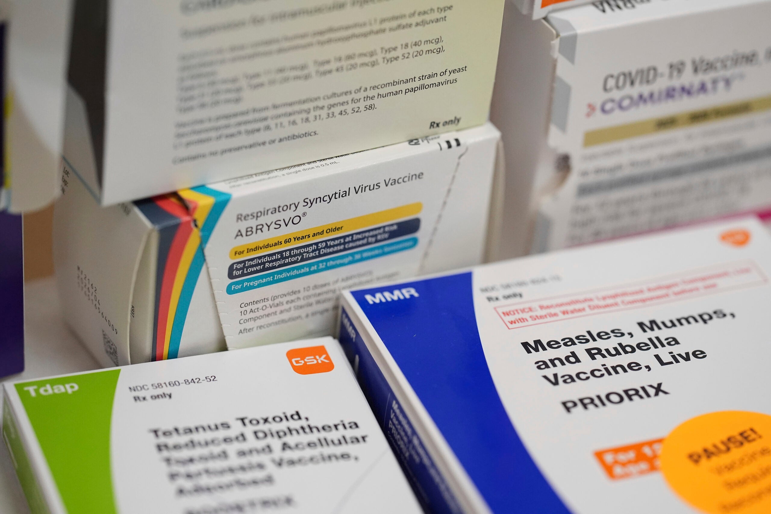 Boxes of various vaccines, including Tdap, MMR, COVID-19, and RSV, are stacked closely together, displaying their labels and dosage information.