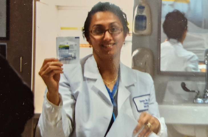 Nurse in a white coat holds up a medical test packet while standing in a clinical workspace.