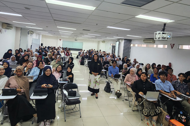 Large classroom of students seated at desks, facing the camera, with an instructor standing in the center aisle.