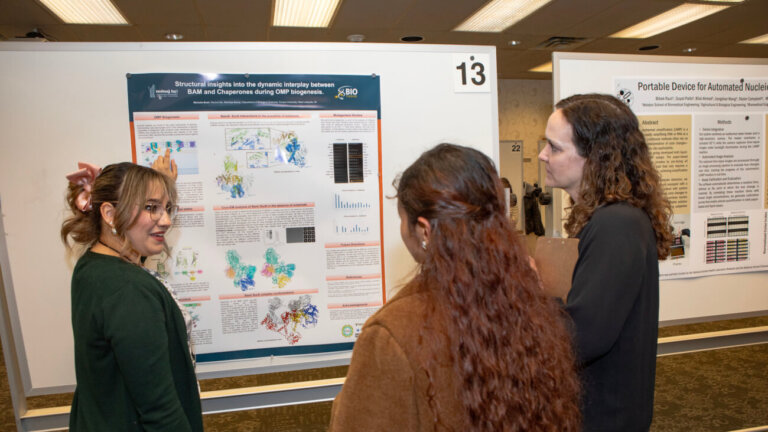 three females stand in front of a research poster. One of the women is wearing a green shirt and appears to be presenting the poster to the other two women.