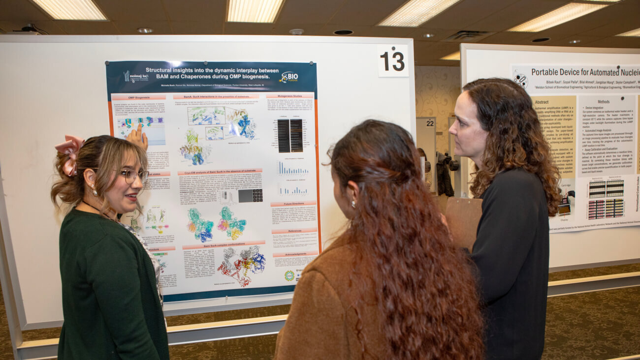 three females stand in front of a research poster. One of the women is wearing a green shirt and appears to be presenting the poster to the other two women.