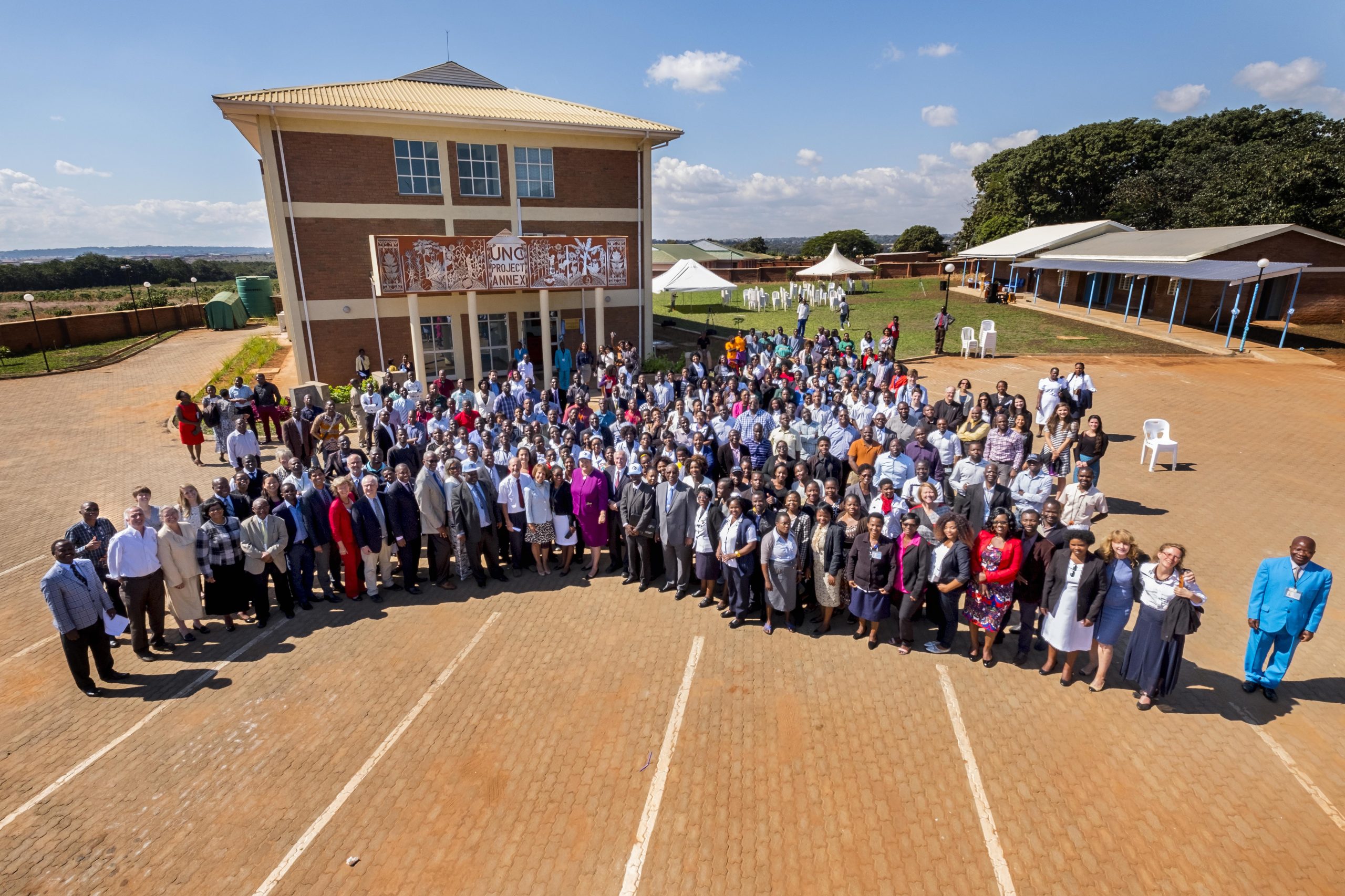 Shows a large group of people standing in front of a building that's set in a field with a sunny blue sky, tents and several other buildings in the background. Myron Cohen stands in front row of the group.