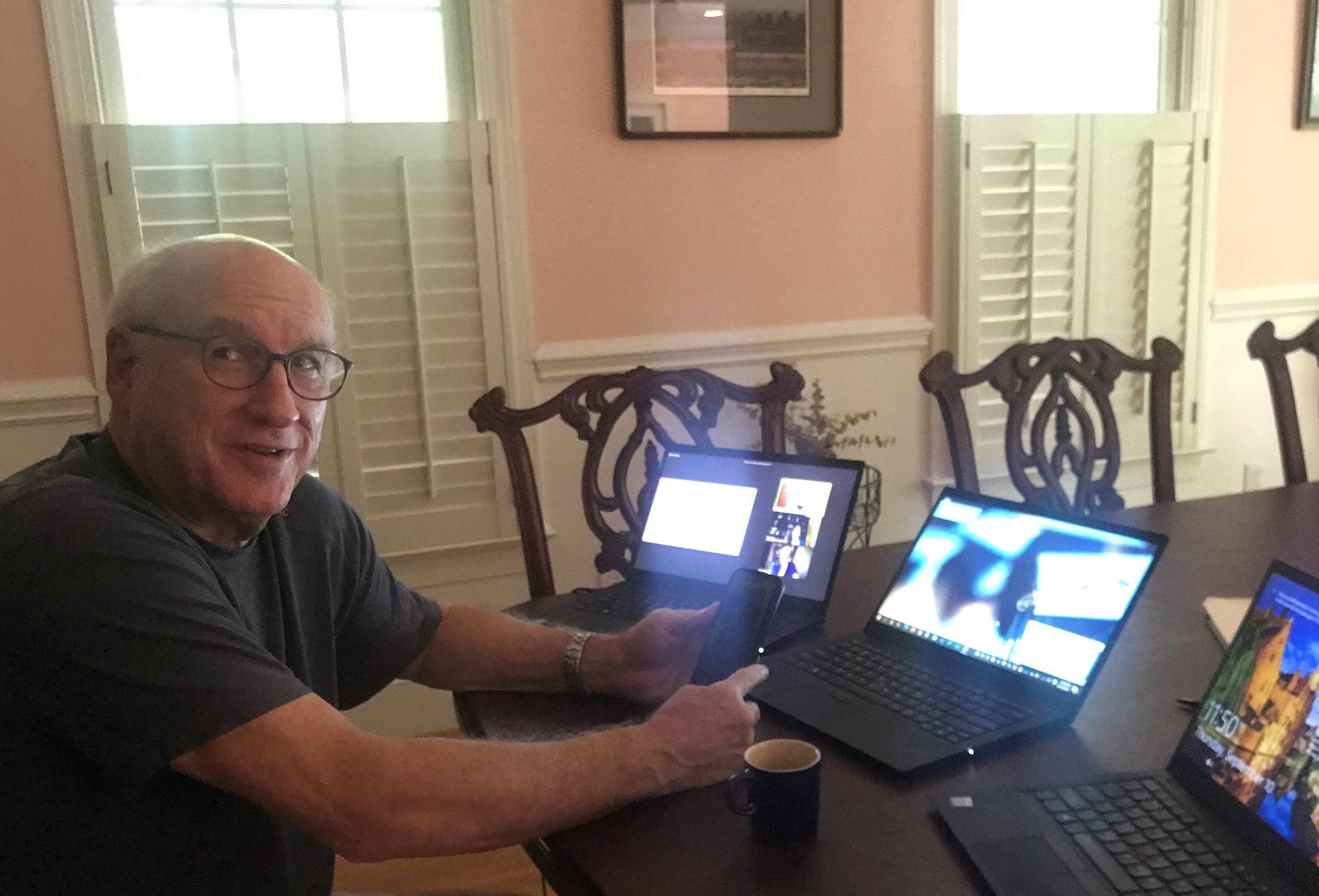 shows myron Cohen in a t shirt sitting at a formal dining table. In front of him are three laptops, all on, one has an active meeting on it. He's also holding a smart phone.