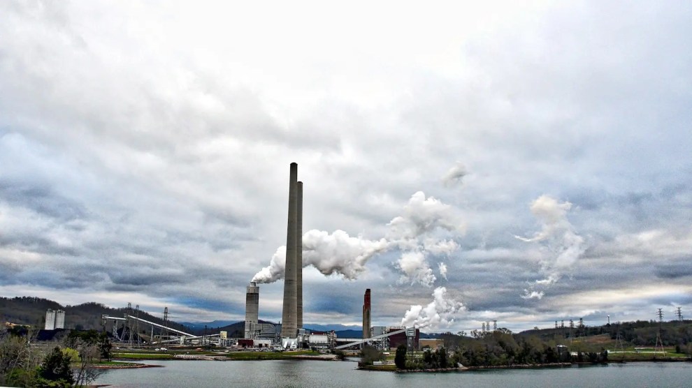 Smoke billows out of smokestacks of a power plant at the edge of a pond with green hills in the background.