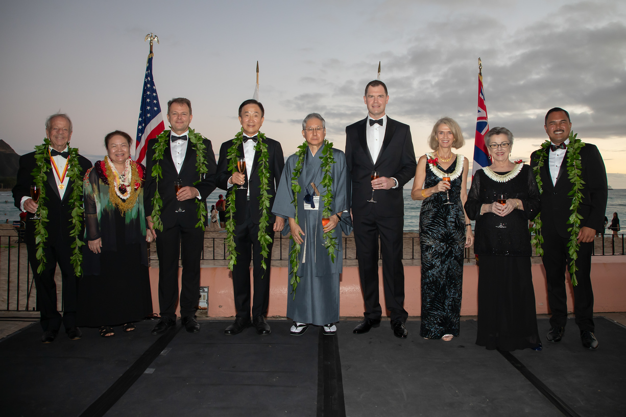 Ladies and gentlemen dressed in festive attire stand in a row, with flags of various countries behind them.