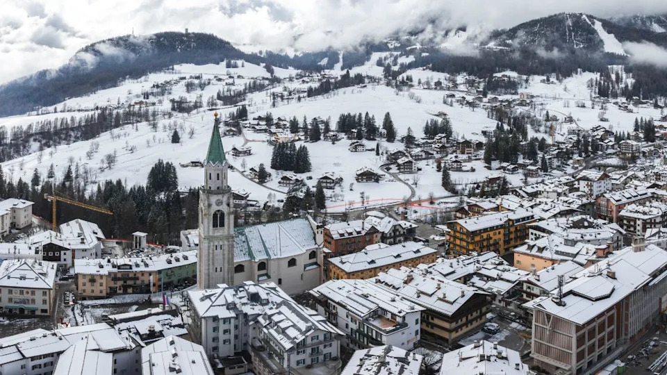 Aerial view of Cortina d'Ampezzo, Italy, featuring the Basilica Minore dei Santi Filippo e Giacomo, snow covered rooftops, and misty Dolomite peaks.