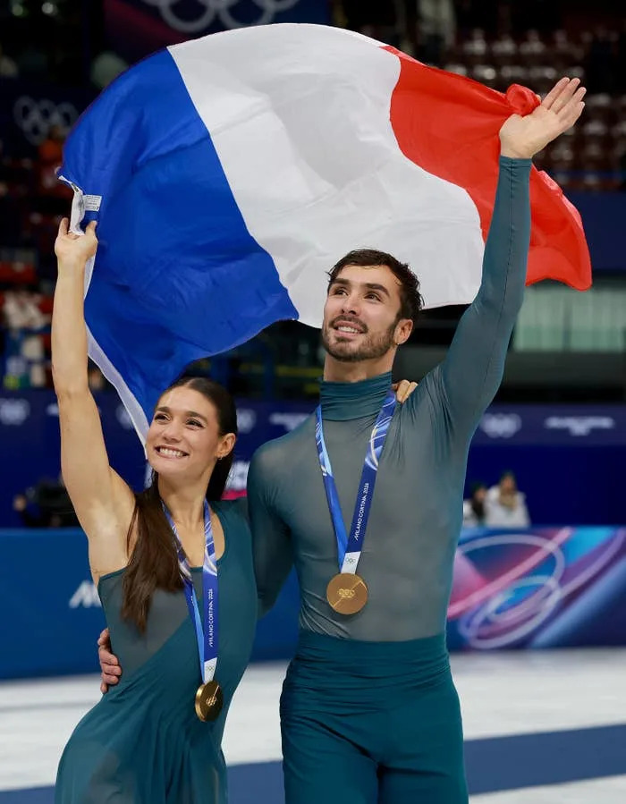 Two athletes celebrating with medals, holding a French flag. Both dressed in elegant, fitted outfits