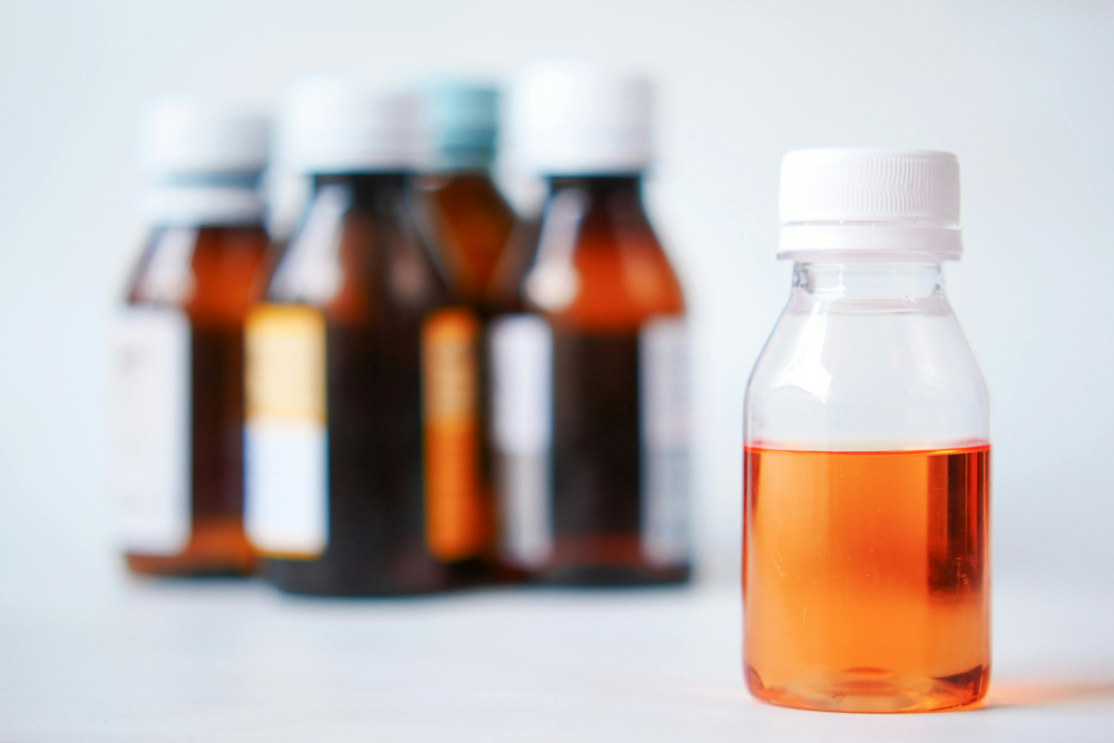 A clear plastic bottle containing orange liquid medicine stands in sharp focus in the foreground, with several amber glass medicine bottles blurred in the background.