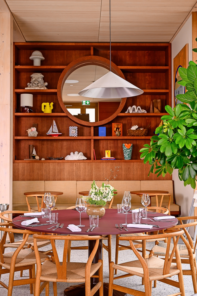 A dining room at Auberge Port-Gitana in Geneva, Switzerland, featuring a round table set with glasses and white napkins, surrounded by wooden chairs, in front of a wooden shelf adorned with decorative objects and a round mirror.