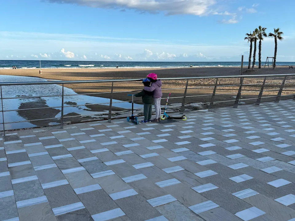 Two children standing next to scooters look out at the ocean.