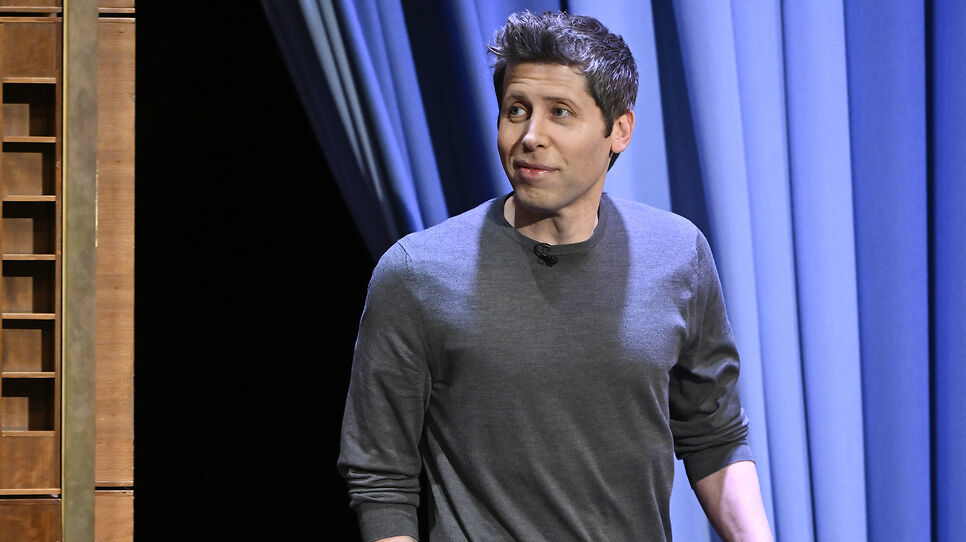 A man in a gray sweater stands on a stage with blue curtains, looking to the side.