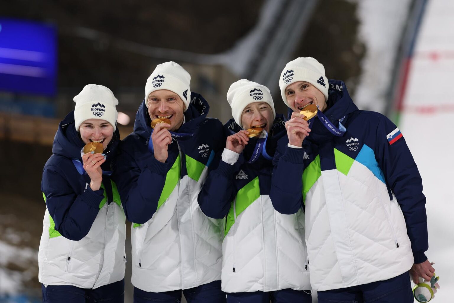 Three athletes in winter gear embrace on a snowy slope with Olympic branding in the background, surrounded by spectators.