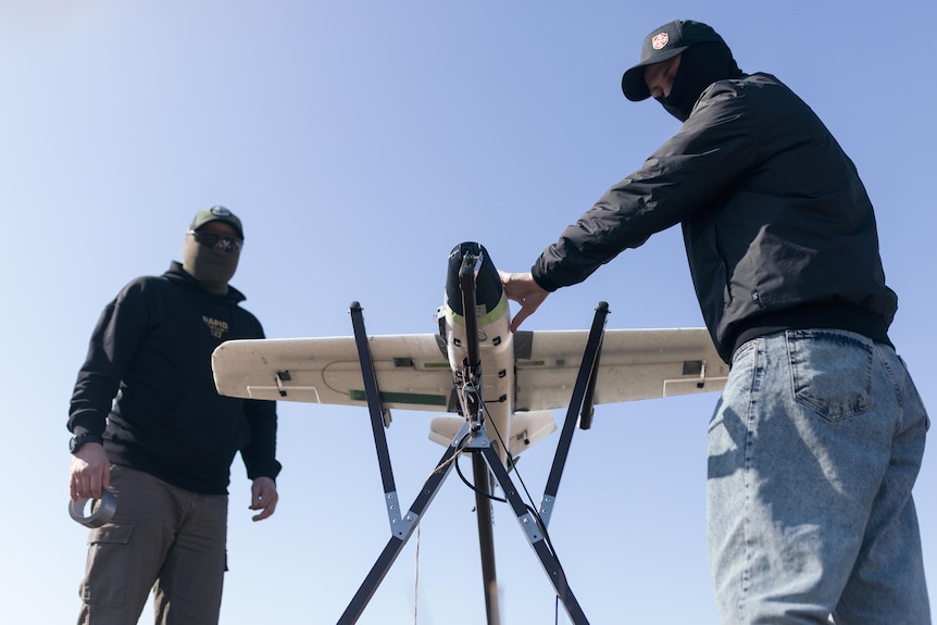 Two men in jeans, jackets and face coverings prepare a small, white, winged drone.